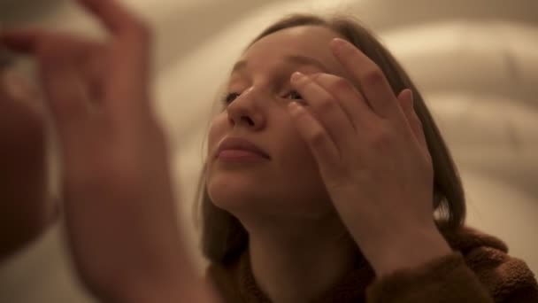 Mooie lichte bruine haired meisje is het toepassen van rimpel crème op ooglid in de spiegel kijken. Handheld beeldmateriaal — Stockvideo