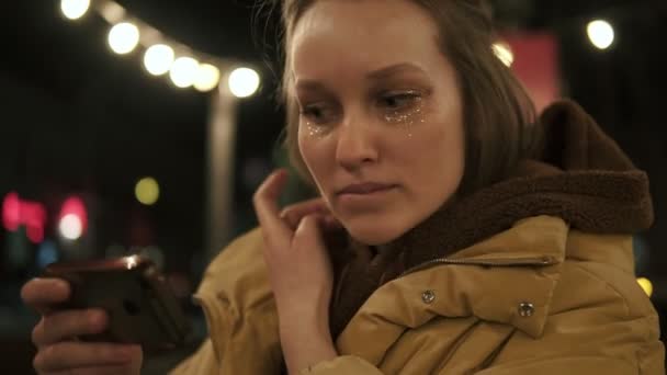 La fille se tient debout dans la rue en regardant dans le petit miroir entraver ses cheveux clairs. Elle scintille sur son visage. Soirée, lampadaires — Video