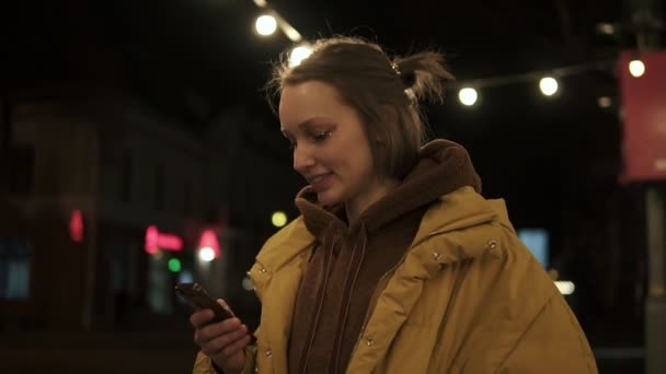 Mooi meisje in het midden van een donkere straat met lichten kijkt naar de telefoon en glimlacht. Gele vacht. Zijaanzicht — Stockvideo