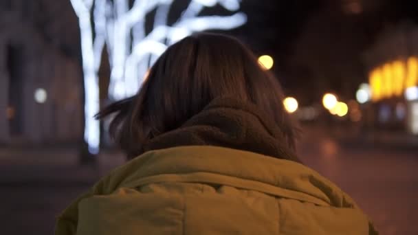 Hermosa chica tierna está caminando por la ciudad de noche en abrigo. Se da vuelta y sonríe. Tiro de seguimiento — Vídeos de Stock