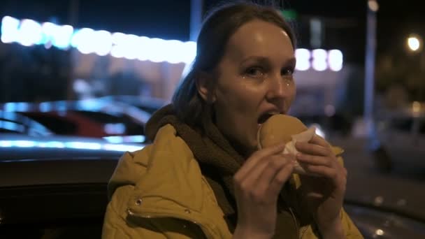 Feliz chica sonriente con una chispa en su cara comiendo hamburguesa en una calle nocturna. Imágenes de mano — Vídeos de Stock