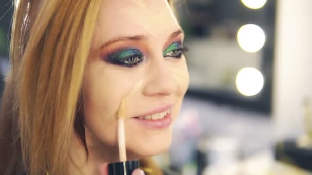 Visagist εφαρμόζοντας το υγρό make up σε νέους χαμογελαστό πρόσωπο γυναικείο χρησιμοποιώντας ραβδί. Επαγγελματικό μακιγιάζ στο σαλόνι. Πλάγια όψη — Αρχείο Βίντεο