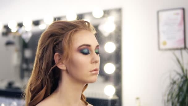 Prachtige Kaukasische model met lang gedraaide haar en kameleon ogen make-up. Stylist make-up met spray afwerking. Salon spiegel op achtergrond — Stockvideo