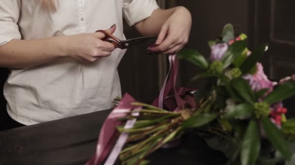 Una chica con una camisa blanca corta cintas largas de color rosa y cintas anchas de guipur de color púrpura para decorar un ramo de flores en una mesa. De cerca. — Vídeos de Stock