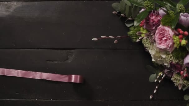 Дві шовкові рожеві стрічки розкидані по темній дерев'яній поверхні, красивий букет квітів в рамці. Повільний рух — стокове відео