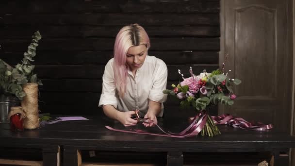 Žena květinářství dokončí složení kytice, uřízne tipy hedvábné stuhy s nůžkami. Nadšen z její práce. Čelní pohled. Dřevěné interiérové pozadí — Stock video