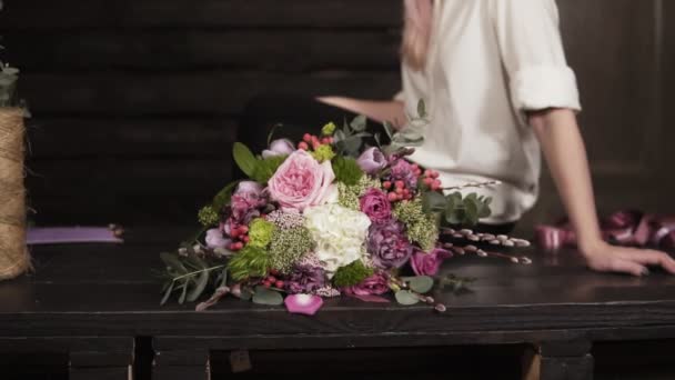 Hermosa grabación en cámara lenta de un increíble ramo hecho por un florista profesional. Chica seductora y sonriente con camisa blanca está sentada en la mesa. Estudio — Vídeo de stock