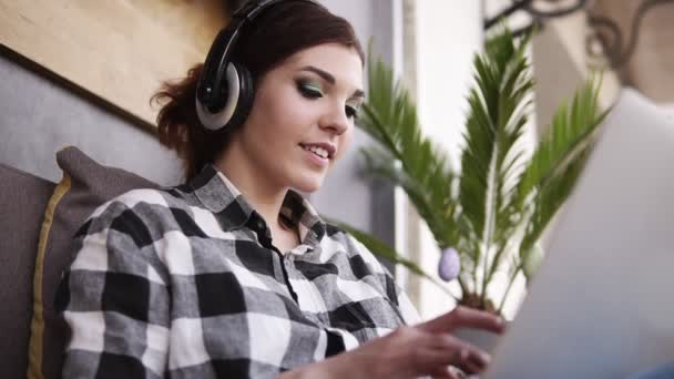 Primer plano de material de archivo de hermosa, porrista, chica morena en camisa a cuadros. Chica feliz, charlando con alguien usando un portátil. Auriculares. En interiores — Vídeos de Stock