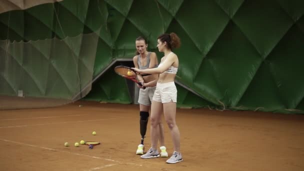 Красивая женщина-тренер с протезом на ноге показывает своему подопечному нюансы удара мяча в теннисе, стоящем позади нее. Спортивные девушки — стоковое видео