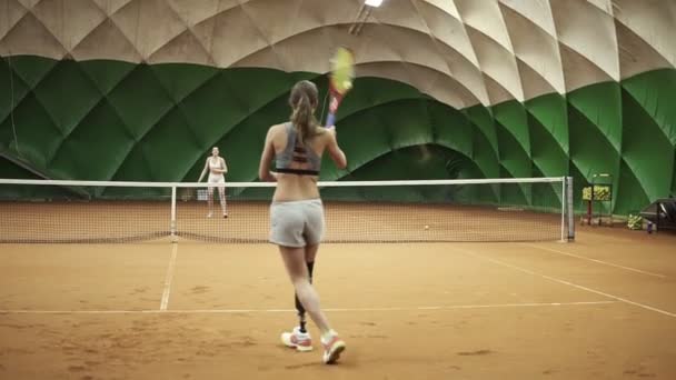 Imágenes de dos chicas atléticas jugando al tenis en una pista de tenis cubierta. Mujer en el marco con prótesis de pierna — Vídeos de Stock