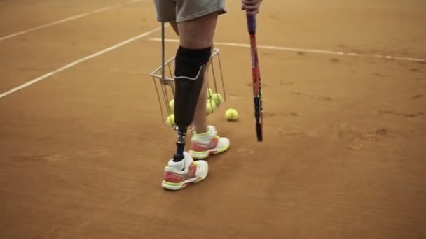Seguimiento de imágenes de una chica deportiva con una prótesis en el pie derecho recogiendo pelotas de tenis con la cesta. Jugador de tenis. Vista trasera — Vídeo de stock