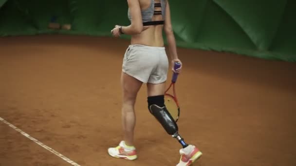 Спортивная девушка-инвалид берет мяч из корзины, идет к середине и делает сильный выстрел теннисной ракеткой. В помещении — стоковое видео
