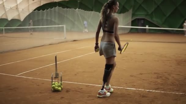 Dos chicas hermosas practicando. La chica discapacitada en pantalones cortos toma la pelota de la canasta y hace el primer servicio de la pelota. Pista de tenis cubierta. Vista trasera — Vídeos de Stock