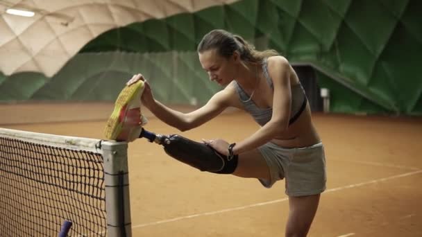 Ein schlankes behindertes Mädchen streckt vor dem Spiel ihr verletztes Bein auf dem Tennisnetz aus. Prothese. drinnen — Stockvideo