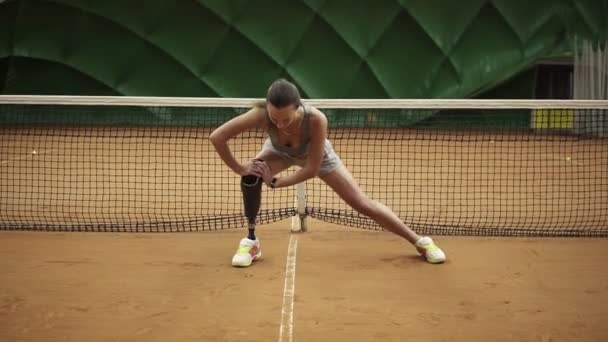 Ein schönes Mädchen dehnt ihre Beinmuskeln auf einem überdachten Tennisplatz. Behindert. Tennisnetz zurück. Frontansicht — Stockvideo