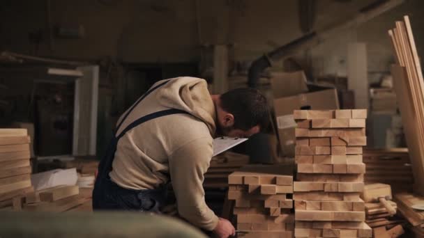 Человек в столярной мастерской с табличкой и ручкой считает количество деревянных блоков. Джойнтер в рабочей одежде. Вид спереди — стоковое видео