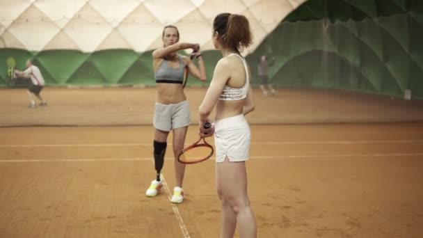 Een mooie, atletisch ongeldige meisje leert de beginner van het juiste traject van een hit in tennis racket. Andere spelers op de achtergrond. Slow motion — Stockvideo