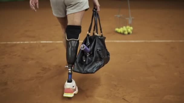 Lächelnde Athletin kommt zum Tennistraining, zückt einen Schläger und geht auf den Platz. Beinprothese. Zeitlupe — Stockvideo