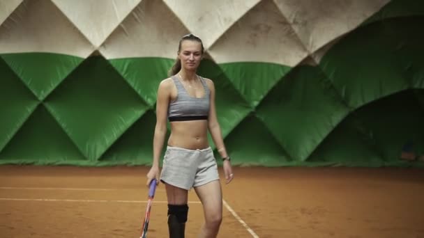 Jovem de cabelos compridos em sportswear está andando no campo de ténis interior com raquete na mão. Tenda verde atrás. Movimento lento — Vídeo de Stock