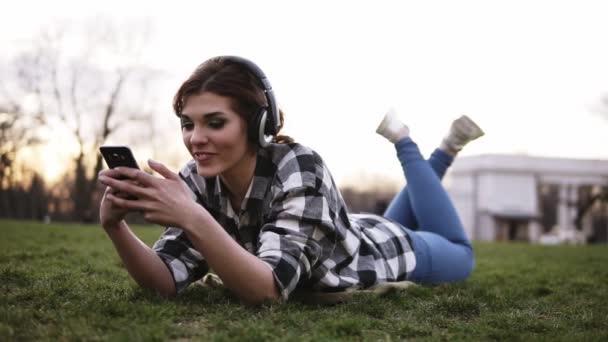 Счастливая молодая женщина слушает музыку на смартфоне с наушниками, лежащими на зеленой траве в парке. Девушка развлекается, используя свой мобильный телефон на открытом воздухе — стоковое видео