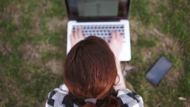 Apuntando imágenes de una chica con una cola de pelo se sienta en el suelo en un parque, impresiones en una computadora portátil borrosa. El móvil está en la hierba cerca. Disparando de arriba hacia abajo — Vídeos de Stock