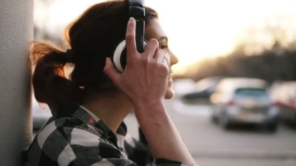 Fondo borroso brillante al aire libre. Vista lateral de una hermosa chica que se pone los auriculares y relajarse. Escucha la música. Apóyate en la pared. Sonriendo. — Vídeo de stock