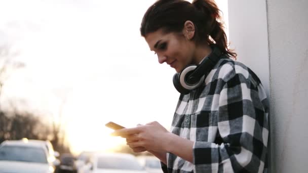 Mulher bonita está de pé perto da coluna branca, segurando móvel em suas mãos e digitação. Auscultadores. O sol está brilhando — Vídeo de Stock