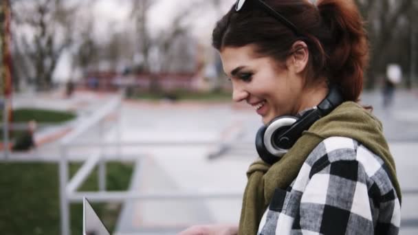 Cheerful young girl with a laptop in her hands walking through the park and typing at the same time. Laughing and having fun. Blurred background — Stock Video