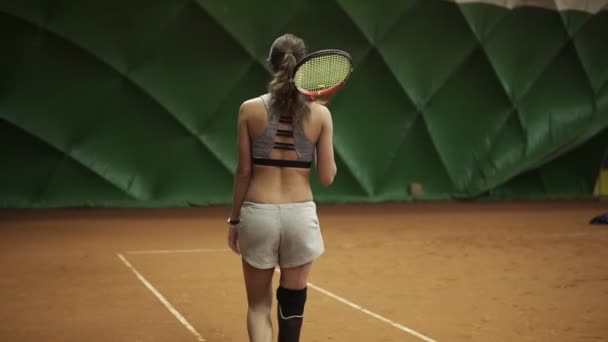 Una chica alta con una prótesis está caminando a lo largo de una pista de tenis con una raqueta en el hombro. Pantalones cortos grises y sujetador. Pista de tenis cubierta. Vista trasera — Vídeo de stock