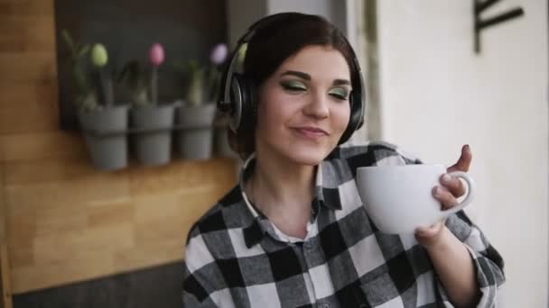 Brunette dívka tančí v potěšení s šálkem čaje a sluchátka poslouchat oblíbenou hudbu. Zpomalený pohyb. Uvnitř — Stock video