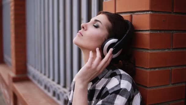 A mulher ouve pensativamente a música nos fones de ouvido, apoiando-se na parede da cerca na rua. Os meus olhos estão fechados. Faz as pazes. Fechar — Vídeo de Stock