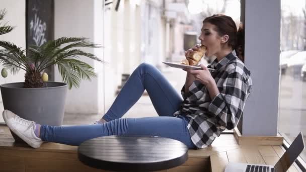 Seitenansicht eines jungen, hübschen Mädchens, das in einem Café auf einem Holzsitz neben den hellen Fenstern sitzt. das Mädchen isst ein Croissant. kaut sie und schließt die Augen. die Zeit genießen — Stockvideo