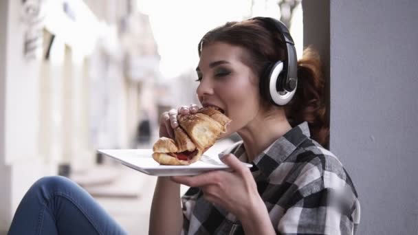 Güzel, genç bir kadın büyük kulaklıklar penceresinde yanında oturan. Müzik ve bir kruvasan yemek dinlemek. Plaka onun kollarında tutarak. Mutlu, gülümseyen. Ağır çekim — Stok video