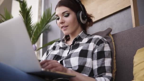 Tiroteio de baixo de uma morena encantadora sentada em um sofá. Menina em fones de ouvido digitando no laptop, sorrindo. Comunicação — Vídeo de Stock