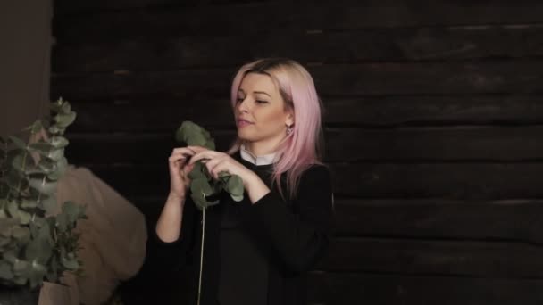 Una chica atractiva y feliz con el pelo rosado quita un puñado de hojas verdes de una rama y juguetonamente lanza a la cámara — Vídeo de stock
