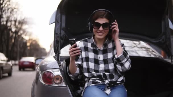 Morena sonriente escuchar la música en los auriculares mientras está sentado en el maletero de los coches abiertos entre la carretera. Teléfono celular. Buen humor. — Vídeo de stock