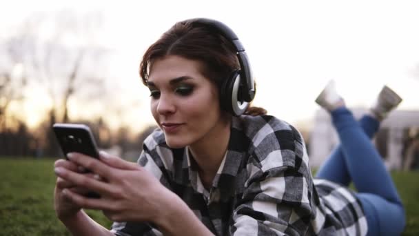 Молодая девушка в стильных наушниках лежит на траве, слушает музыку и болтает в мобильном телефоне. Закрыть — стоковое видео