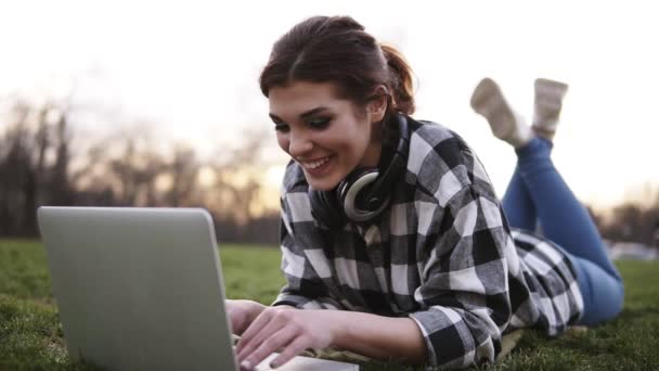 Hermosa chica se encuentra en el césped en el parque. Usa un portátil, charlando. Auriculares en el cuello. Se lo pasa bien en comunicación. Sonrisas, feliz. Día brillante — Vídeos de Stock