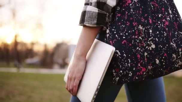 Uitzicht vanaf de kant van het meisje in spijkerbroek wandelen door het park met een gekleurde tas en een laptop in de hand. Close-up — Stockvideo