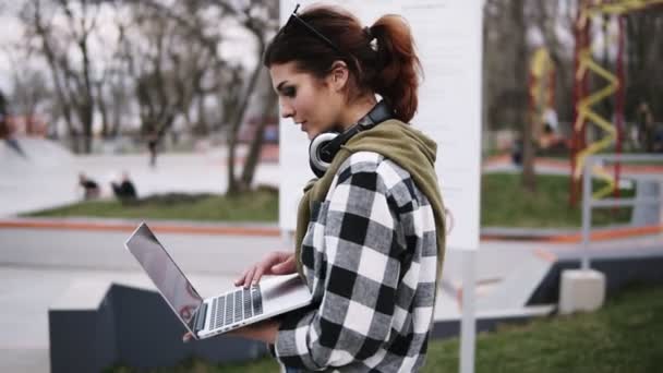 Uma menina bonita e serena caminha pelo parque. Com uma mão ela segura um laptop e envia mensagens com alguém. Olha para a moda. Movimento lento — Vídeo de Stock