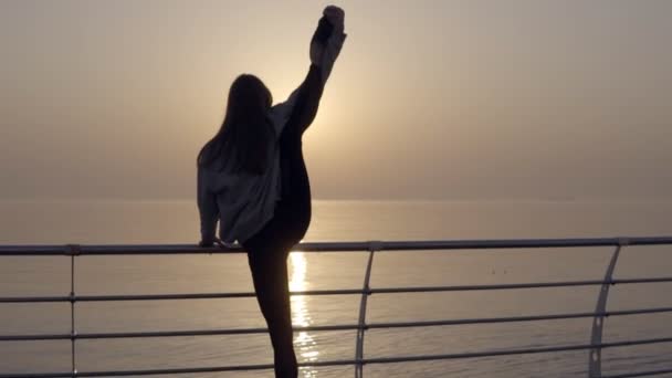 Mujer delgada con piernas largas haciendo estiramiento al amanecer junto al mar, cuerpo perfecto. Estira la pierna sobre la cabeza. Vista trasera — Vídeos de Stock