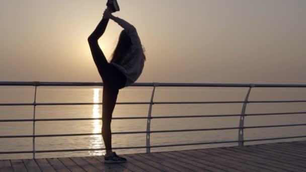 Langhaariges Mädchen streckt ihr Bein mit Blick auf das Meer. Sonnenaufgang. Sportbekleidung, Leggings, Kapuzenpullis. Abenddämmerung — Stockvideo