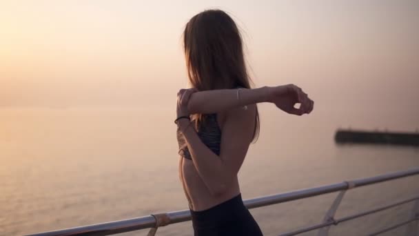 Primer plano de una hermosa joven en un sujetador deportivo estira los músculos de su brazo. Rubia de pelo largo. El sol brilla sobre el océano. Vida deportiva — Vídeos de Stock