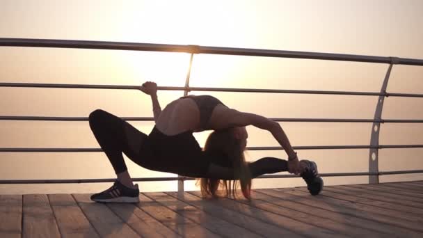 Long-haired girl in sportswear stretches the muscles of the legs, makes lunges. Leans on the crossbar. The morning sun shines on the background. Plasticity — Stock Video