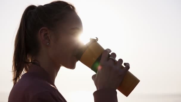 Feche de perto uma jovem linda e sonhadora. A rapariga está a gostar do momento. Bebidas de uma caneca de esportes. Respiração. Sol está brilhando no fundo — Vídeo de Stock