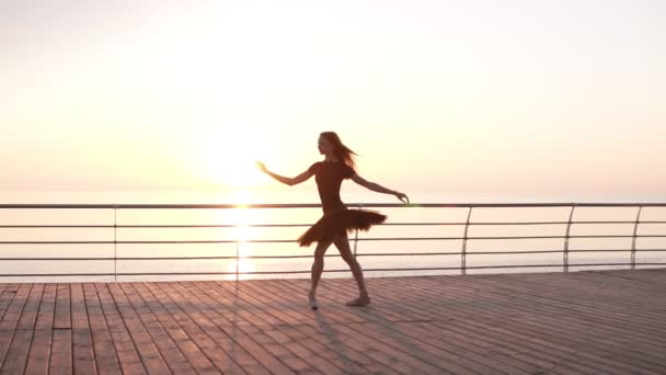 Baletka v černých baletní sukně a bodem na náspu nad moře při západu slunce nebo oceánu. Na lyžích balerína, praktikování klasické cvičení. Zpomalený pohyb — Stock video