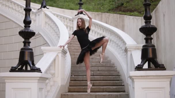 Vue de face d'un ballet gracieux. Jeune ballerine blonde aux cheveux qui fait de la ficelle verticale et pose debout sur un début d'escalier de rue en tutu noir. En plein jour. Mouvement lent — Video