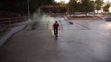 Sarı duman bombası arka tekerleklerde yanarken, boş kaykay parkında pratik yapan beyaz bir BMX sürücüsü. Adam numara yapıyor, bisiklete biniyor ve eğleniyor.