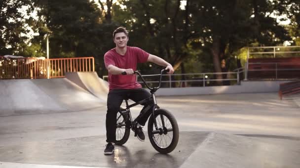 Un joven con un BMX asientos en bicicleta en su bicicleta con rampa, parque de skate en el fondo. Retrato de BMX rider. Cultura callejera. Lente destellos en el fondo. Steadicam tiro — Vídeo de stock