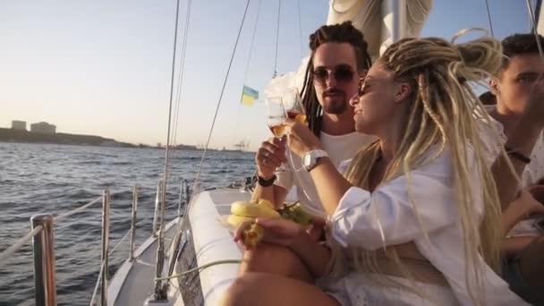 Casal elegante com dreadlocks em roupas brancas e óculos de sol sentado abraçando a borda do iate sorrindo, clinking com óculos champangne. Amante casal passar tempo juntos no iate — Vídeo de Stock